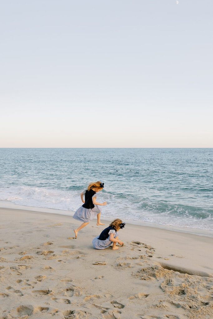 Sisters in matching navy dresses with big navy bows in their hair are playing on the beach in the sand along the shoreline. 