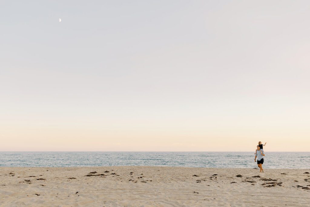 The ocean and beach are the focus here from left to right with a man holding his daughter on his shoulders far off in the distance walking in the sand away from the water. 