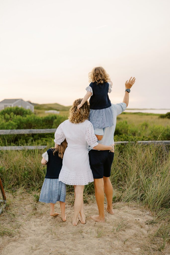 A family of four are standing clustered together on the sand. Mom's arm is around dad's waist to her right and her young daughter's shoulder to her left. Dad is holding the toddler girl on his shoulders waving at the ocean, while the toddler girl is clutching mom's curls. 