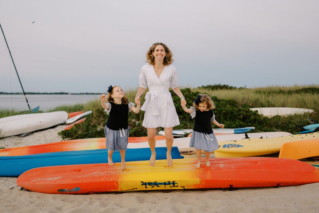 A curly-haired mom wearing a white cotton wrap dress is holding both of her daughter's hands while preparing for all three to jump off of an overturned orange and yellow kayak they are standing on top of. 