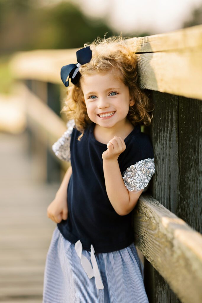 A blue-eyed, curly-haired brunette toddler girl with a big navy bow in her hair is leaning on against the post of an old wooden bridge with a huge smile on her face. 