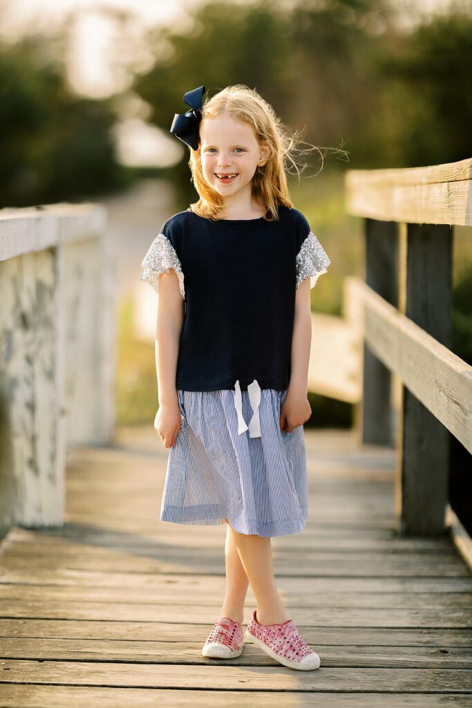 Strawberry blonde young girl is standing on a old wooden bridge wearing a blue dress and pink Native slip on shoes with a big navy bow in her hair. She is missing her two front teeth and smiling big. 