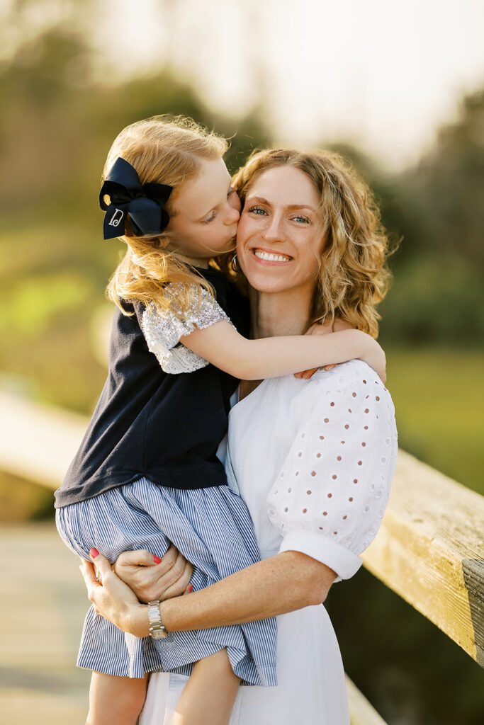 Mom wearing a white dress with eyelet elbow-length sleeves and curly hair is smiling at the camera and holding her daughter wearing a blue dress with a big navy bow in her strawberry blonde hair and is kissing her mom's cheek. 