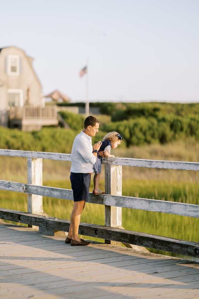 Dad in navy shorts and light blue linen button-down is supporting his curly-haired daughter as she stands on the railing of an old wooden bridge overlooking the marsh below. 