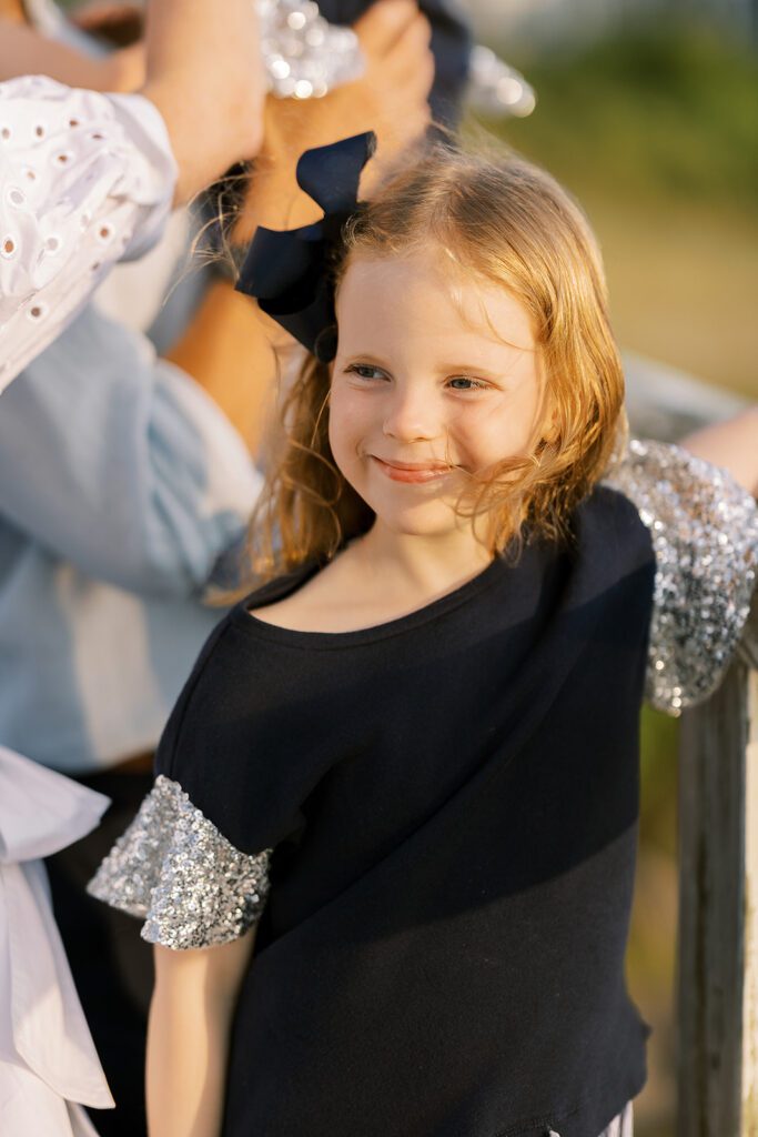 A strawberry-blonde young girl in a navy top with short, sparkling flutter sleeve and big navy bow holding her hair off to the side is grinning off into the distance. 