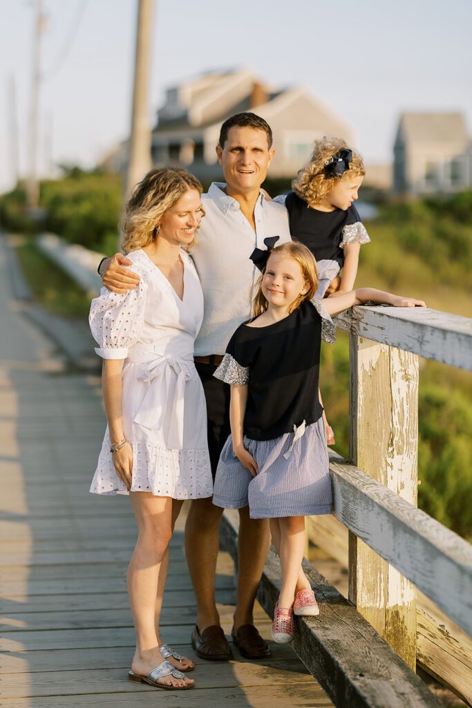 A family of four, mom, dad, and two daughters, are standing along the railing of an old wooden bridge bathed in sunlight and smiling at one another. 