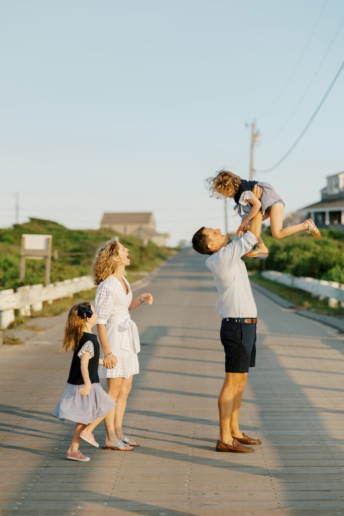 A family of four are standing in the middle of a road leading to the beach. Mom is holding the hand of the eldest daughter as they both are joyous in laughter as the dad standing in front of them raises the youngest daughter high into the air. 