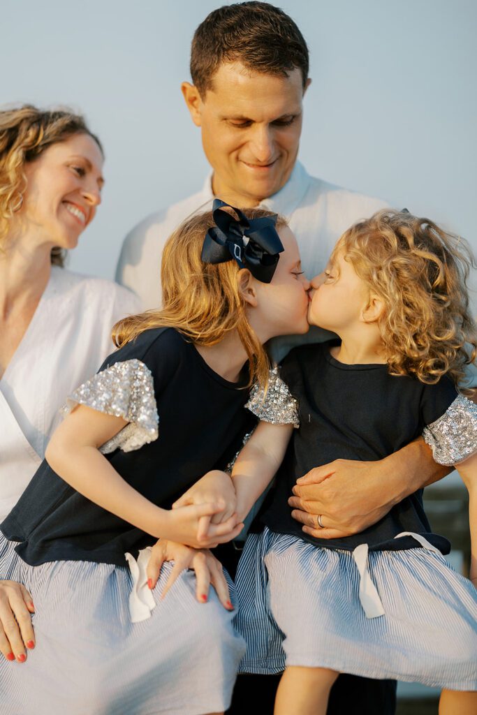 Sisters in matching navy dresses with short, sparkly flutter sleeves are sitting on a railing, holding hands, and giving either other a kiss on the lips as mom and dad stand behind them lovingly smiling upon their daughters.  