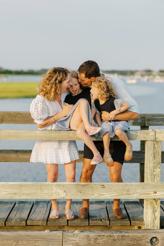 Mom in white eyelet dress with curly shoulder-length hair and dad in navy shorts and brunette hair are holding two daughters as they sit on the top railing of a small wooden bridge. Dad is kissing of his daughter's cheeks while mom and daughters have huge open-mouth smiles on their faces. 