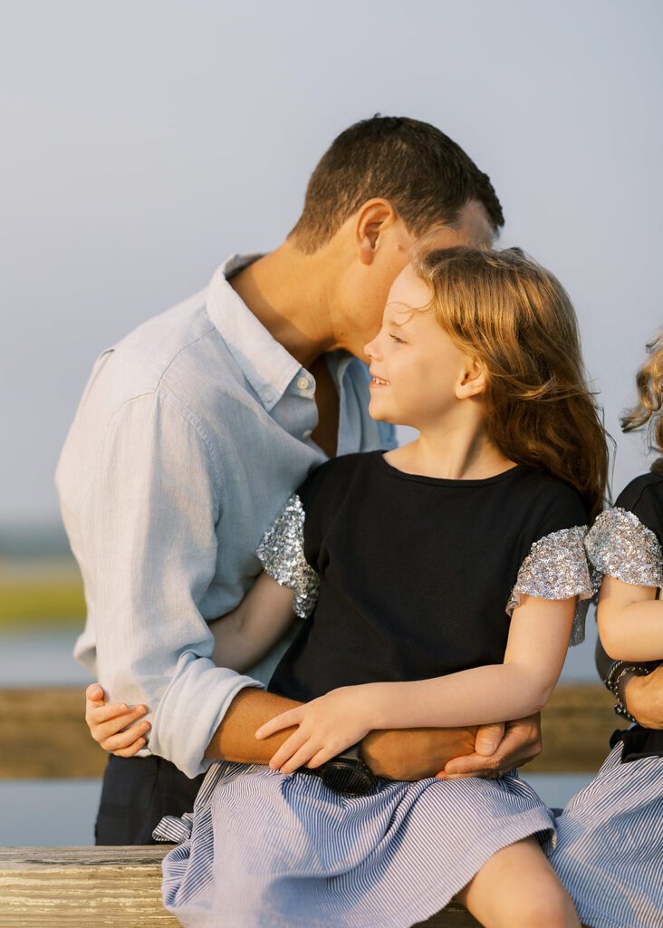 Strawberry blonde-haired young girl is sitting on the top railing of a wooden bridge with her dad standing behind her with his arms around her waist. She is smling off to the side as her dad's face is hidden from view as he whisper's something in her ear. 