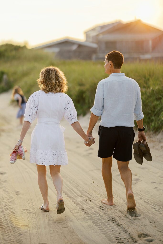 A man and woman are walking hand-in-hand onto the beach. He's wearing navy shorts and a light blue collared shirt, holding his shoes in his hands, while she is wearing a white dress with eyelet detail that hits right above the knee. 