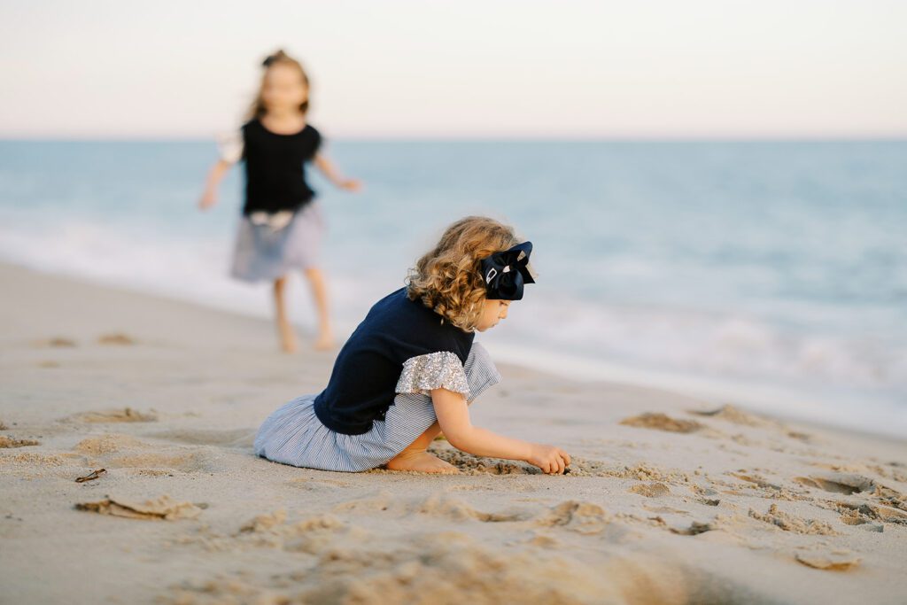 A young toddler girl with brown curls and a big navy bow in her hair is crouching down playing in the sand. Her big sister wearing a matching dress is running towards her in the background though blurry to the eye. 