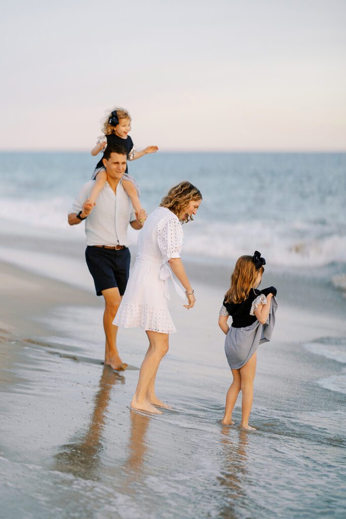 A family of four is standing in the wet sand of the beach as the ocean wave retreats. Dad has the youngest daughter on his shoulders walking towards Mom, who is wearing a white dress and is saying something to her older daughter who is standing right in front of her holding her dress up so not to get it wet. 