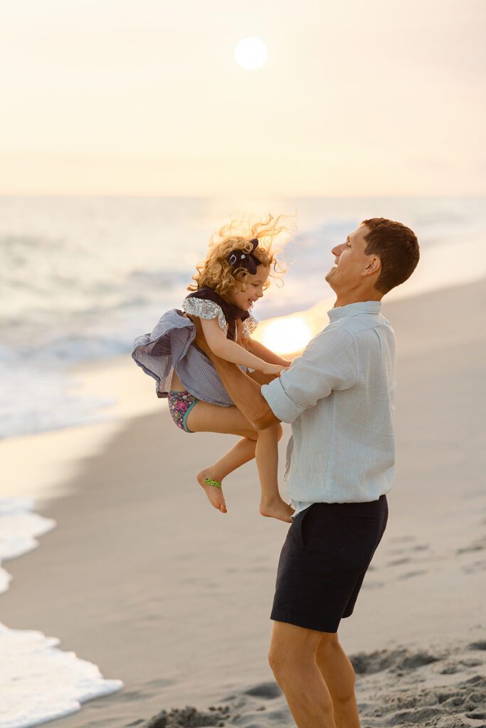 Dad in navy shorts and a light-colored button-down is standing on the beach along the shoreline at sunset and is mid-lift with this toddler daughter. 