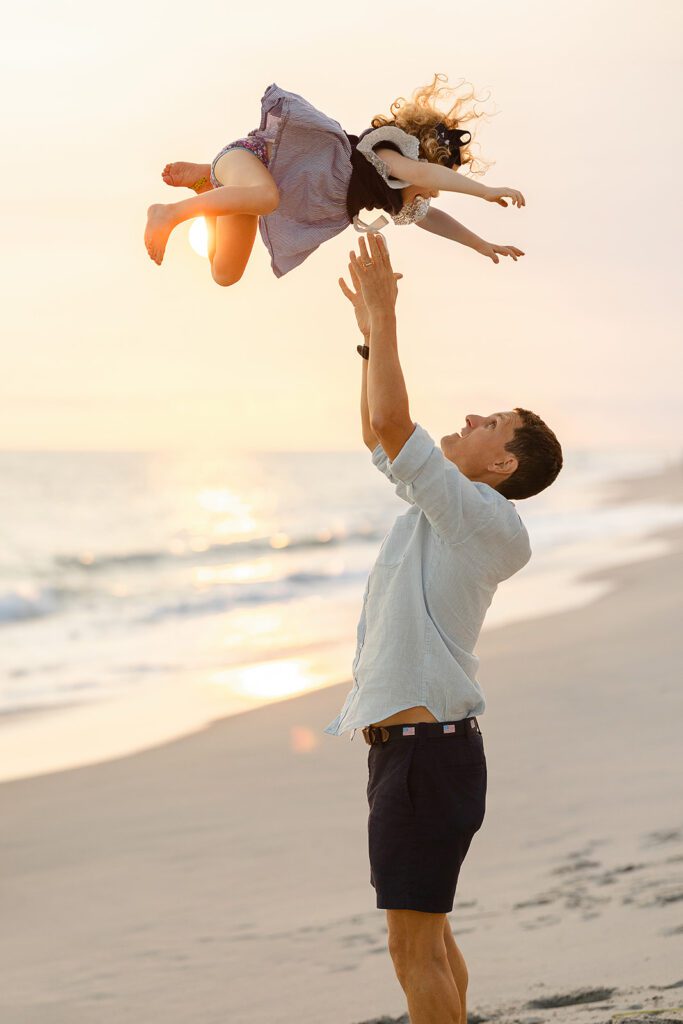 Dad in navy shorts and a light-colored button-down is standing on the beach along the shoreline at sunset having just tossed his curly-haired toddler daughter into the air so it appears like she's flying. 