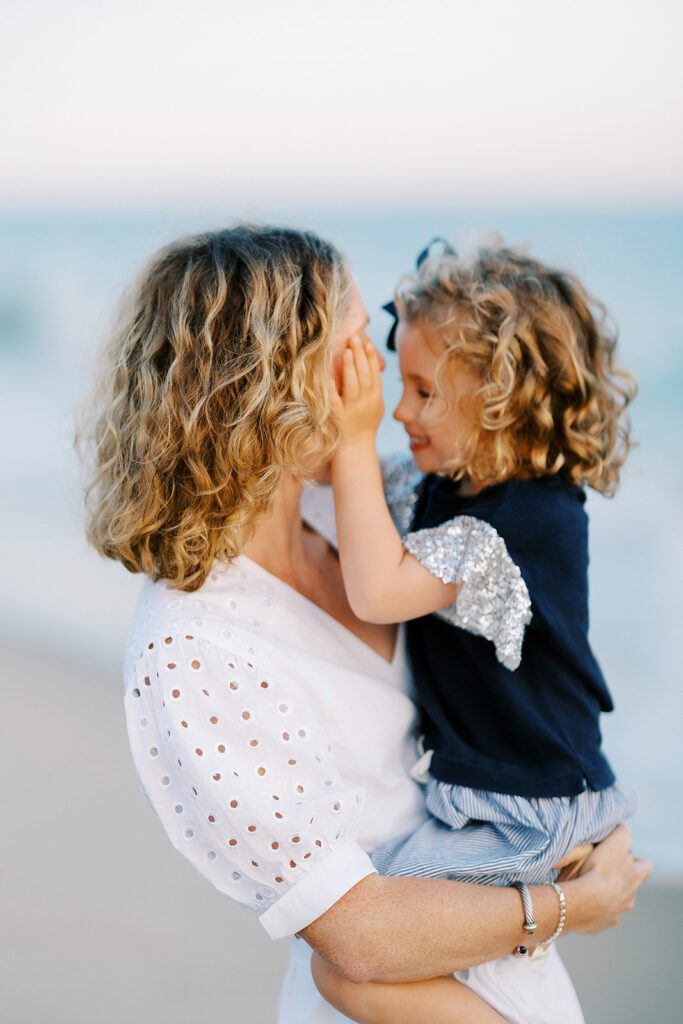 A curly-haired mom in a white short-sleeve dress is holding her toddler daughter with matching curls. The little girl is cupping mom's face and smiling at her. 