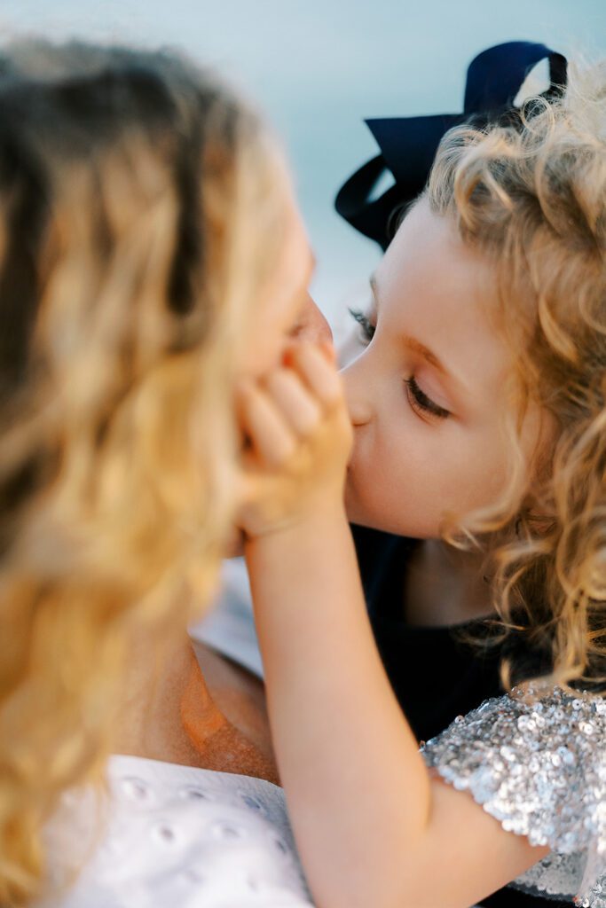 Toddler girl with big navy bow is holding mom's cheeks and giving her a kiss. Mom's face is hidden from view. 