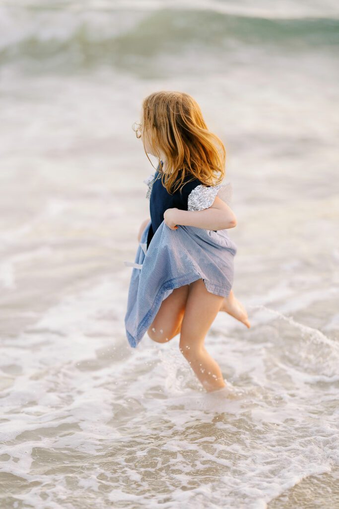 A strawberry-blonde girl is holding her dress up and running through the the shallow ocean waters while looking out to sea. 