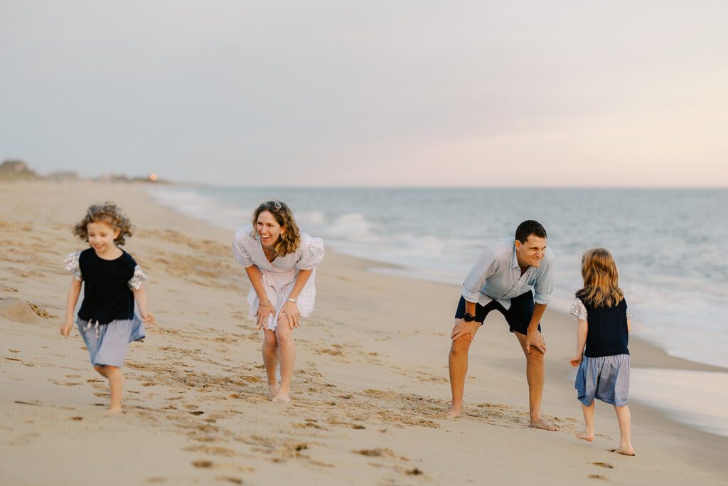 A family of four is on the beach standing by the shoreline. Mom is bend down, hands on her knees, smiling at her curly-haired toddler girl who is smiling and running in the opposite direction. Dad is bending down, hands also on his knees, making a silly smiling face at his older daughter who is walking towards him. 