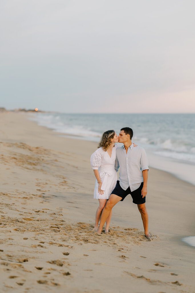 A woman in a white dress and a man in a light blue button down and navy shorts are standing on the beach just in front of the shoreline kissing. 