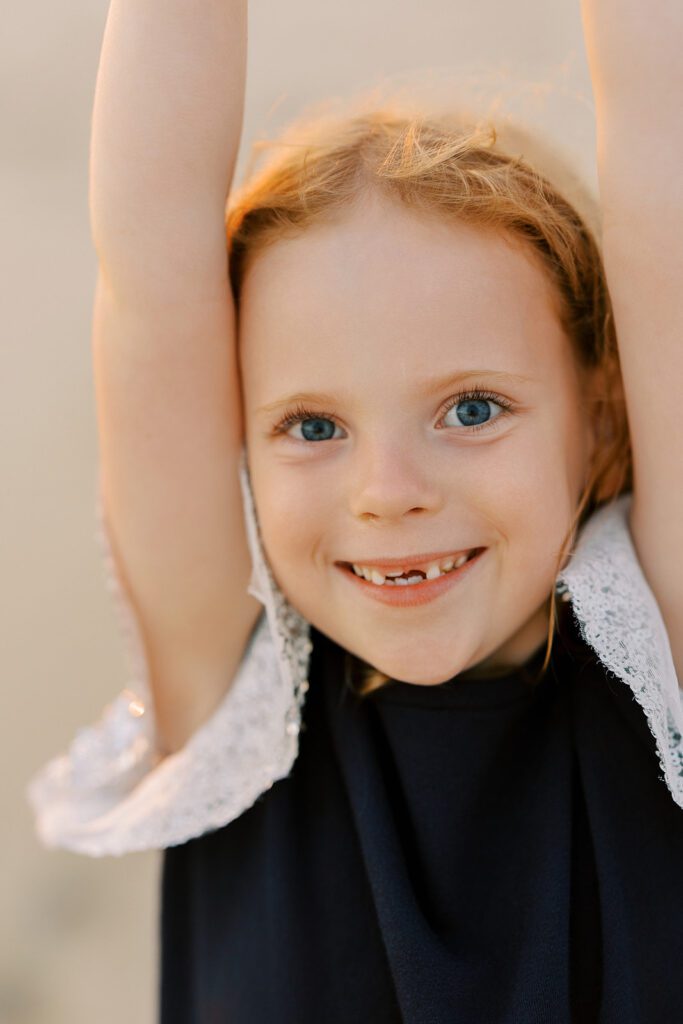 A strawberry-blonde, blue-eyed girl is holding both arms in the air on either side of her head with a big smile on her face showcasing some missing teeth. 