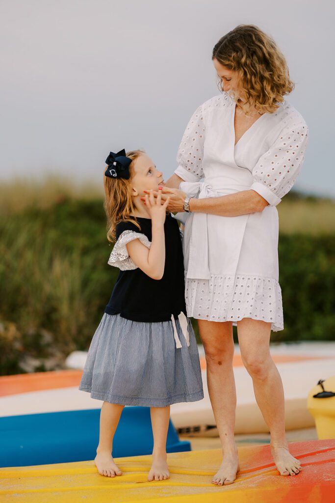 A mom and daughter are standing on top of an orange and yellow overturned kayak on the beach. Mom is wearing a white, wrap, cotton dress and is cupping her daughter's face with her hands and looking down at her. The little girl is smiling up at her mom wearing a navy dress with a big navy bow pinning her strawberry-blonde hair out of her face. 