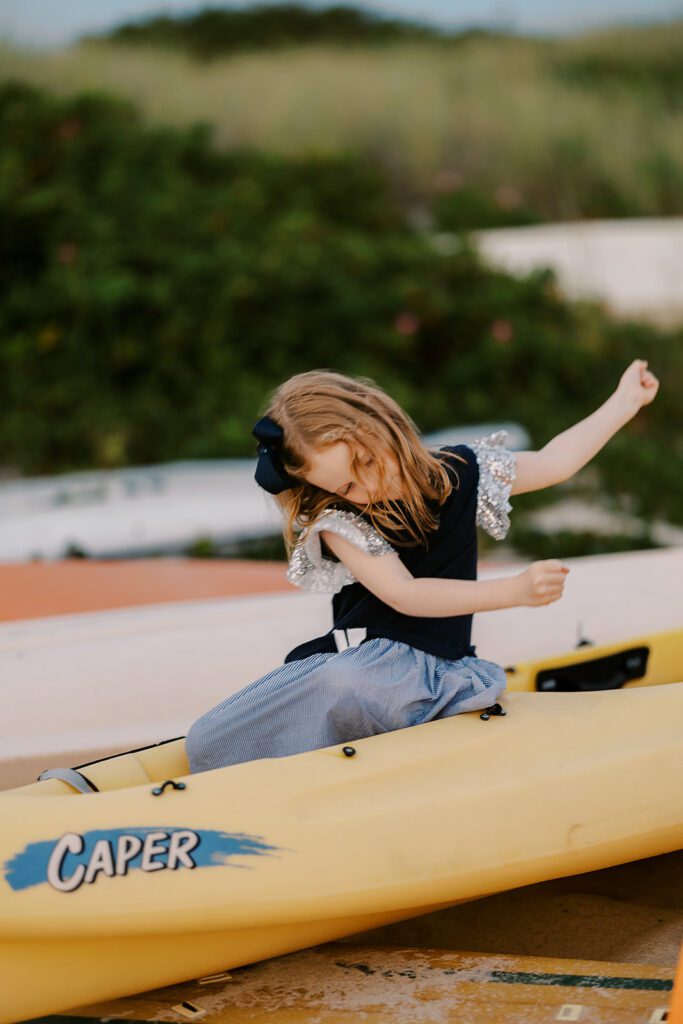 A strawberry-blonde girl with a navy dress and big navy bow is sitting in a yellow kayak on the beach playing. Her arms are in mid-swing to the right as if she's dancing. 