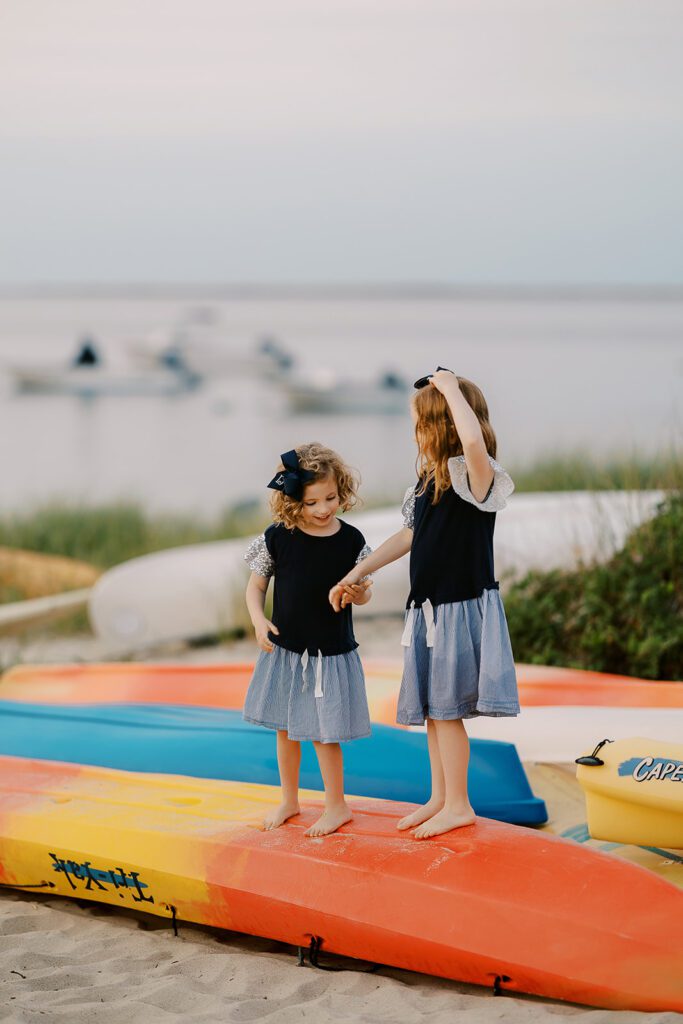 Sisters wearing matching navy dresses with big navy bows are standing on top of an overturned orange and yellow kayak on the beach and holding hands. 