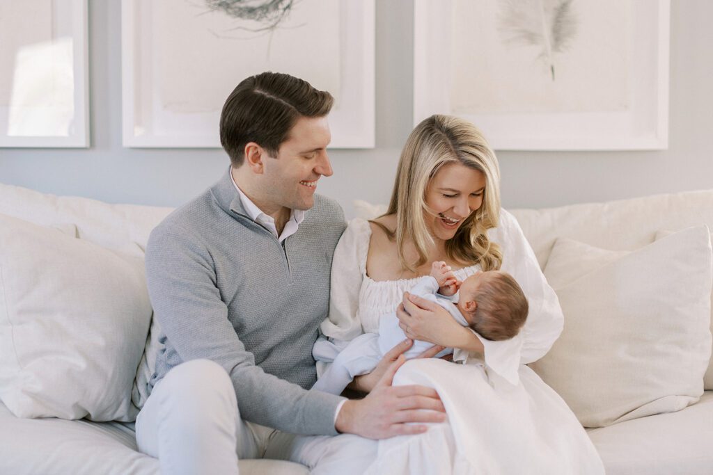 Brunette dad in a gray 1/4 zip pullover is sitting next to his blonde wife in a white linen dress. Mom is holding their brunette haired newborn baby in the nook of her arm and both mom and dad are smiling at their son.