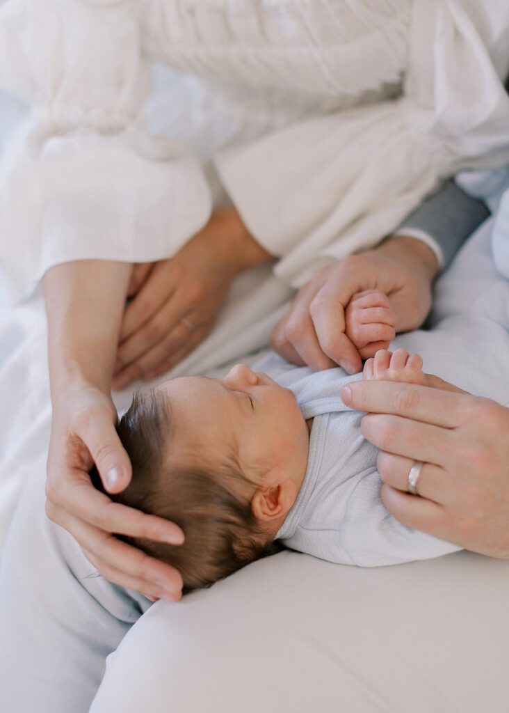 A newborn baby boy with dark brown hair and wearing a light blue cotton crossover onesie is resting on dad's legs. Dad is holding onto the baby's hands and mom is sitting beside them gently resting her hand on the newborn's head.