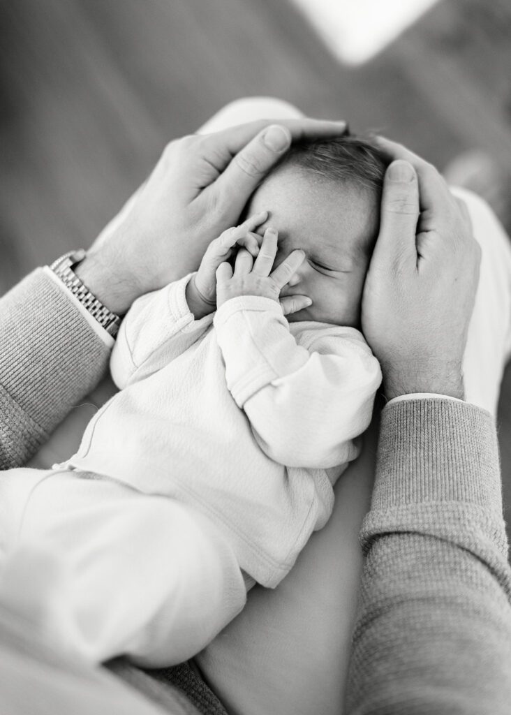 A newborn baby is all scrunched up on his father's legs, with his hands covering his face. Dad is resting both of his hands on either side of his baby's head.