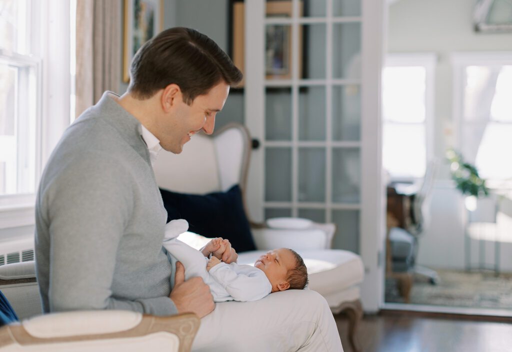 A brunette dad in a gray 1/4 zip sweater and khaki pants is sitting on a chair smiling down at his brunette newborn son who is laying on dad's legs, his head resting at the knees.
