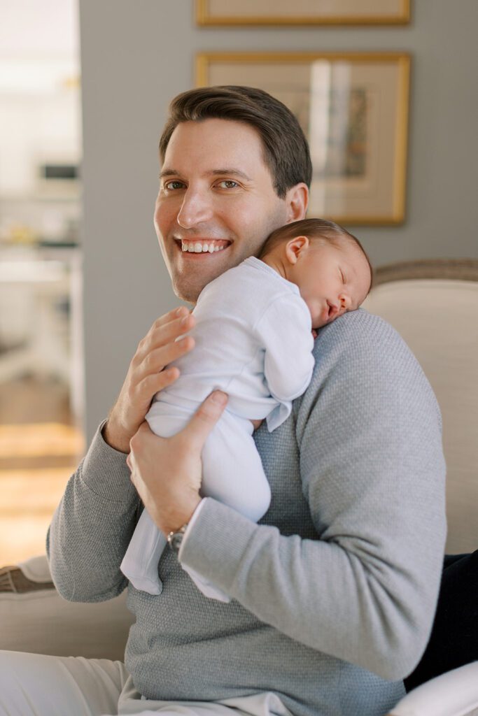 A brunette dad in a gray 1/4 zip sweater is sitting on a chair while holding his newborn sleeping son on his shoulder. Dad is smiling big.
