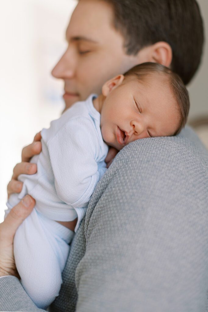 Brunette baby boy is sleeping on his brunette dad's shoulder. Dad is wearing a gray sweater and his face is blurred in the background but you can see his eyes are closed.