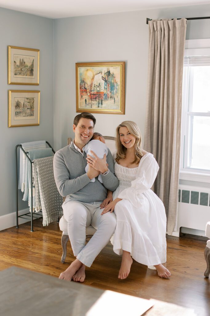 A brunette dad in a gray 1/4 zip sweater is holding his sleeping newborn son on his shoulder. A blonde mom in a white linen dress is sitting right beside dad, her hands resting on his legs, and both are smiling.