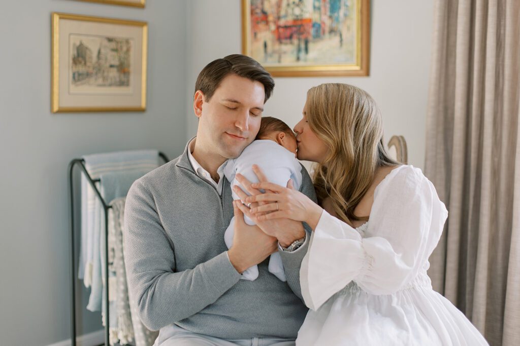 A brunette dad in a gray 1/4 zip sweater is holding his newborn son on his shoulder. Dad's eyes are closed and he's resting his head on the baby. A blonde mom in a white linen dress is sitting right beside dad kissing her newborn son's forehead, her hand resting on her husbands hand who is supporting the baby's bum.
