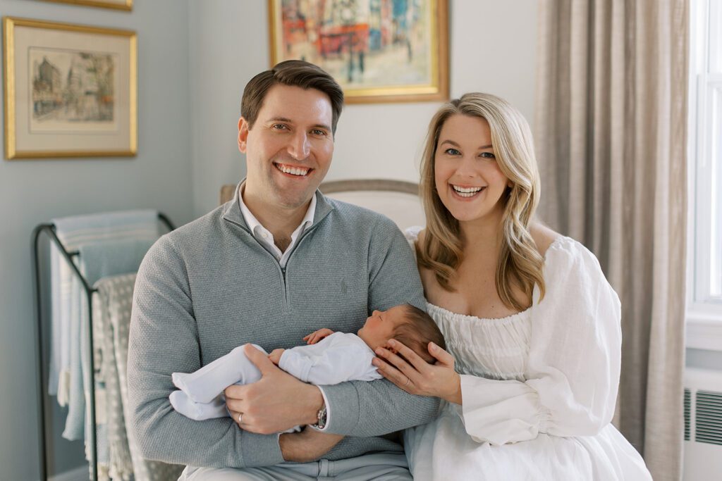 A smiling brunette dad and smiling blonde mom are sitting side by side. Dad is wearing a gray half-zip and cradling his newborn baby, while mom is wearing a white long sleeve dress and resting one hand on the baby's head.