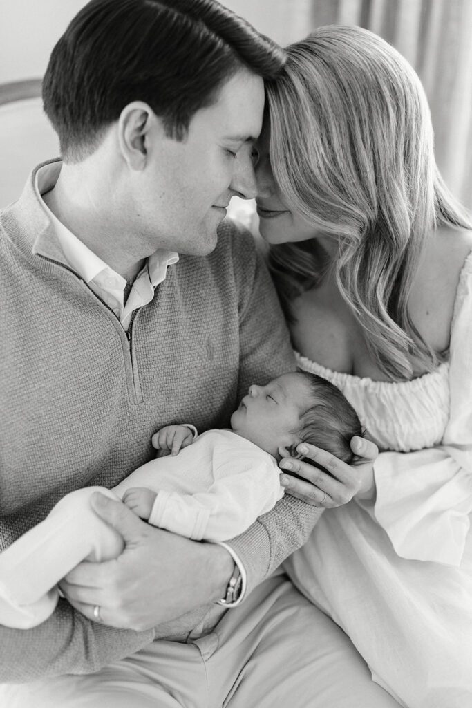 Dad is holding his newborn baby in his arms and sitting next to his wife. They are resting their foreheads against one another with their eyes closed as if soaking in the moment.