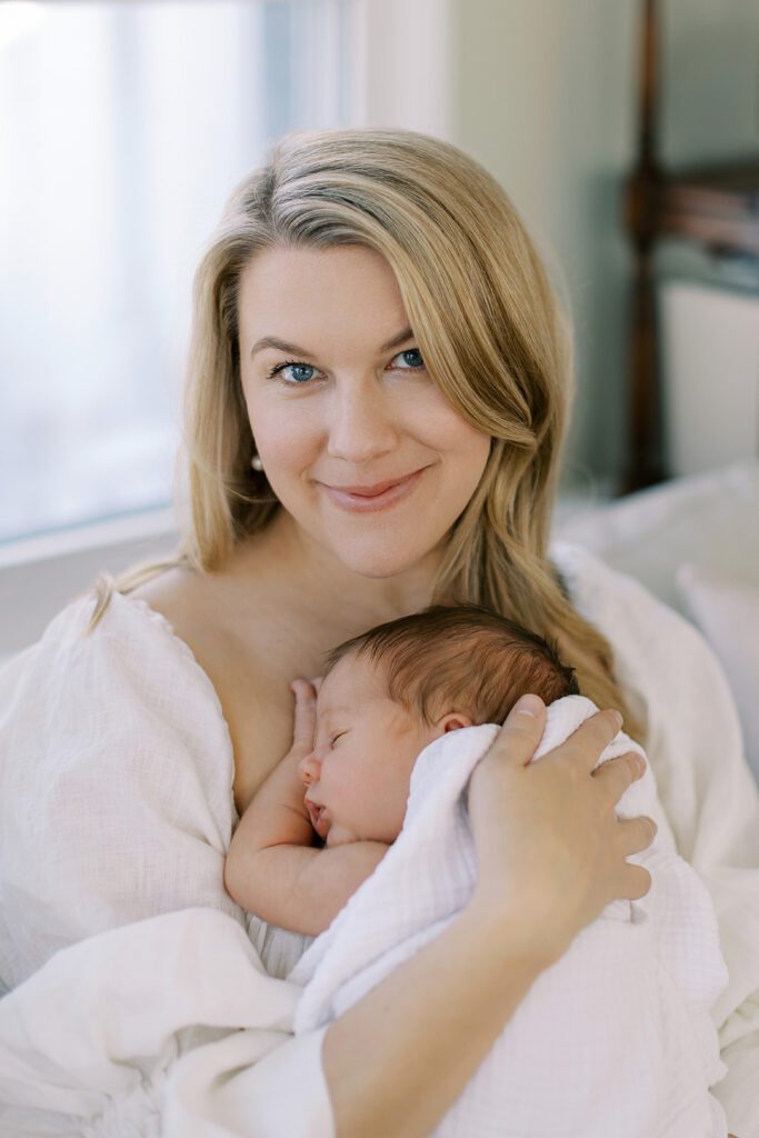 A blonde mom is smiling while holding her swaddled, sleeping brunette newborn baby against her chest.
