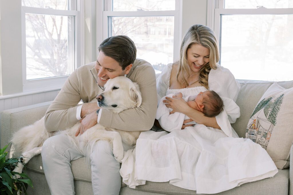 Brunette dad and blonde mom are sitting side by side on their flax colored sofa. Mom is smiling down at her brunette haired baby she is cradling, while a smiling dad is squeezing their golden retriever who is draped over his lap in complete comfort.