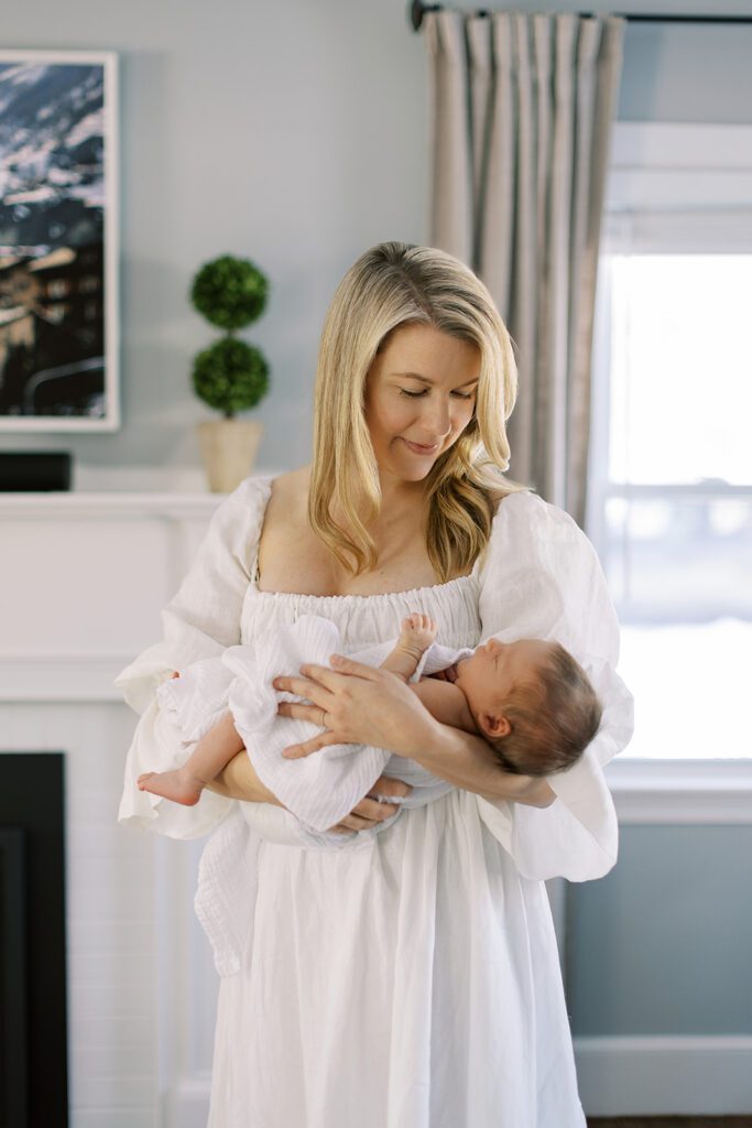 Blonde mom in a white linen dress is standing in front of her fireplace mantle and is smiling down at her newborn baby in the crook of her arm, haphazardly swaddled in a white muslim blanket.