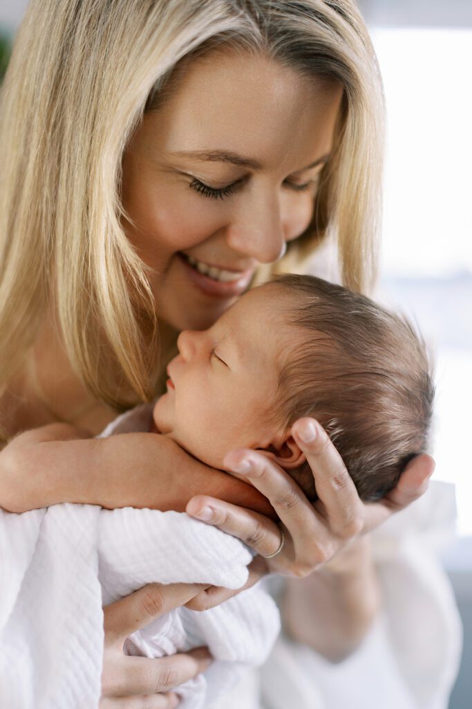 A blonde, smiling mom is holding her brunette newborn baby in her arms up close to her face, about ready to kiss his forehead. The newborn is loosely swaddled in a white muslim blanket with one naked arm poking out, eyes closed.