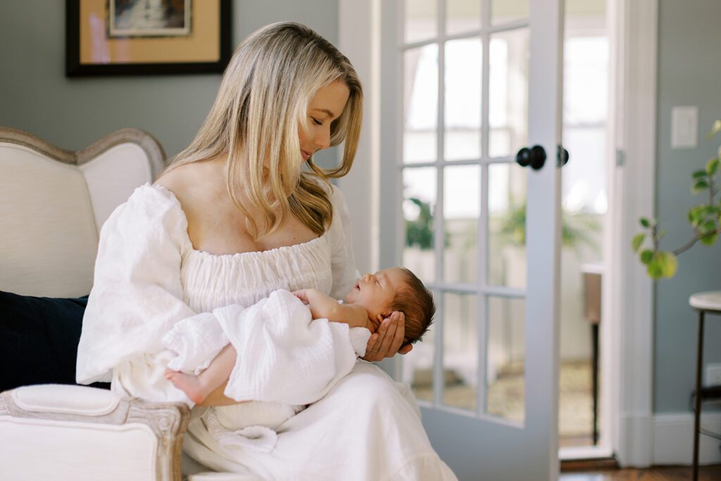 A blonde mom in a white long sleeve linen dress is sitting on a chair with her legs crossed,and cradling her brunette newborn loosely wrapped in a white muslin blanket on her lap.
