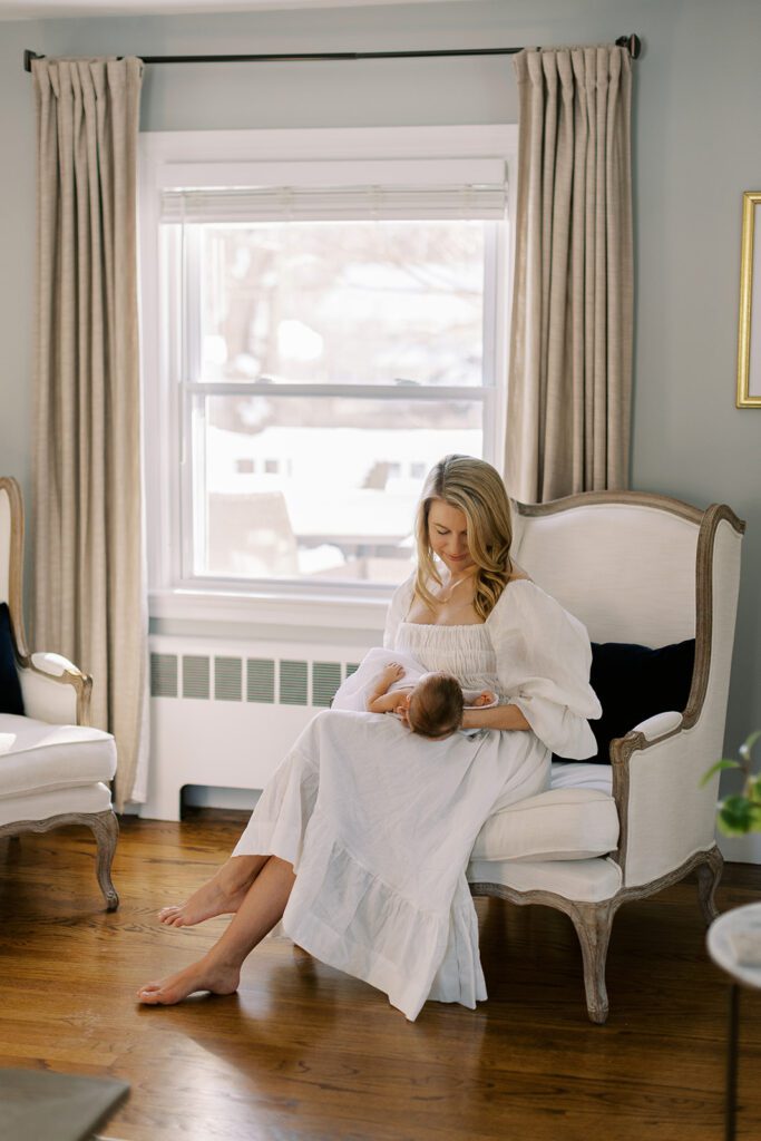 A blonde mom in a white linen dress is sitting on a chair with her legs crossed, gazing down at her dark-haired newborn baby she is resting on her lap.