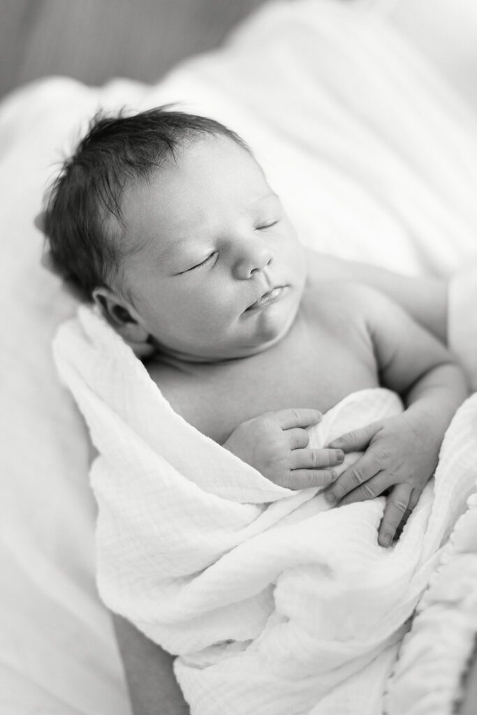 A dark-haired newborn baby is loosely swaddled in a white muslim blanket with one naked shoulder and arm poking out, sleeping.