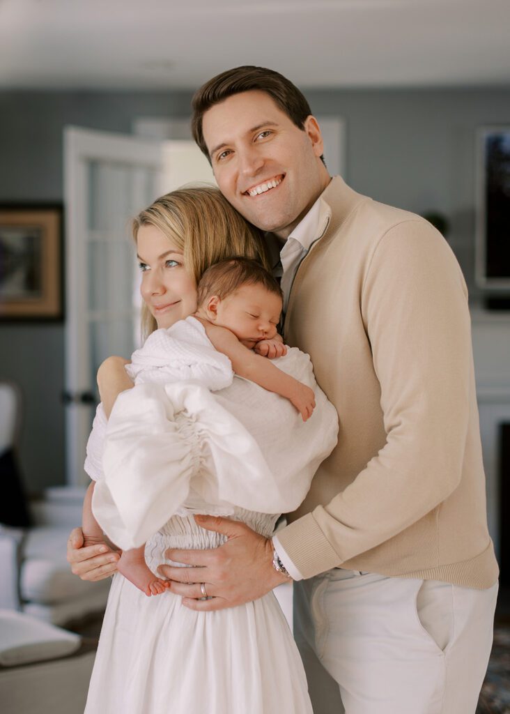 A blonde mom in a white linen dress is holding her swaddled newborn baby on her shoulder, smiling off into the distance, while a brunette Dad is standing behind her, hands on her waist, and smiling big.