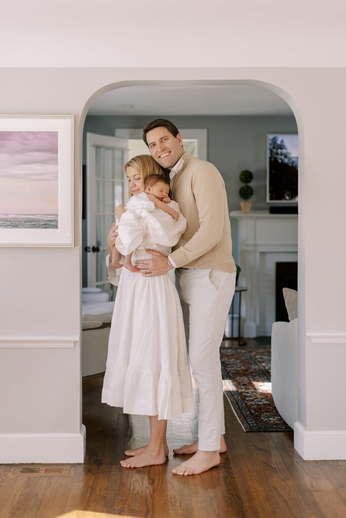 A blonde, barefoot mom in a white linen dress is holding her swaddled newborn baby on her shoulder, her eyes are closed, while a brunette, barefoot Dad in a tan sweater is standing behind her, hands on her waist, and smiling big. They are standing under a curved archway between rooms in their home.