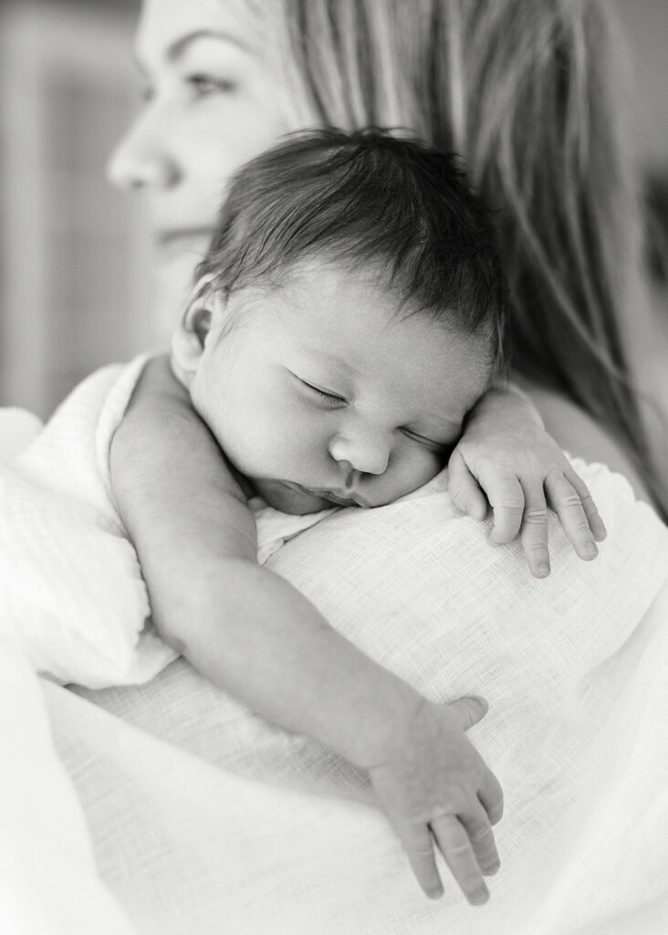 A newborn baby with a full head of hair has one arm flung over mom's arm while resting his head on her shoulder, eyes closed. Mom's face is blurred behind the newborn but you can see she is smiling.