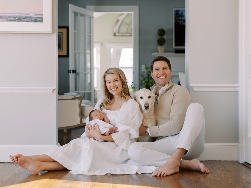 A new family of four are sitting down on the floor together. Mom is wearing a white linen dress with her legs outstretched and feet crossed. She is holding her dark-haired newborn baby boy in her arms. Beside her is her dark-haired husband in a tan 1/4 zip sweater and light pants, smiling big. Their proud golden retreiver is nestled between them.