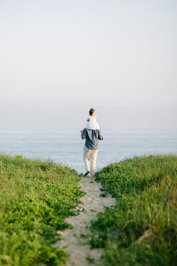 Dad with daughter on shoulders walking down a path toward the ocean.